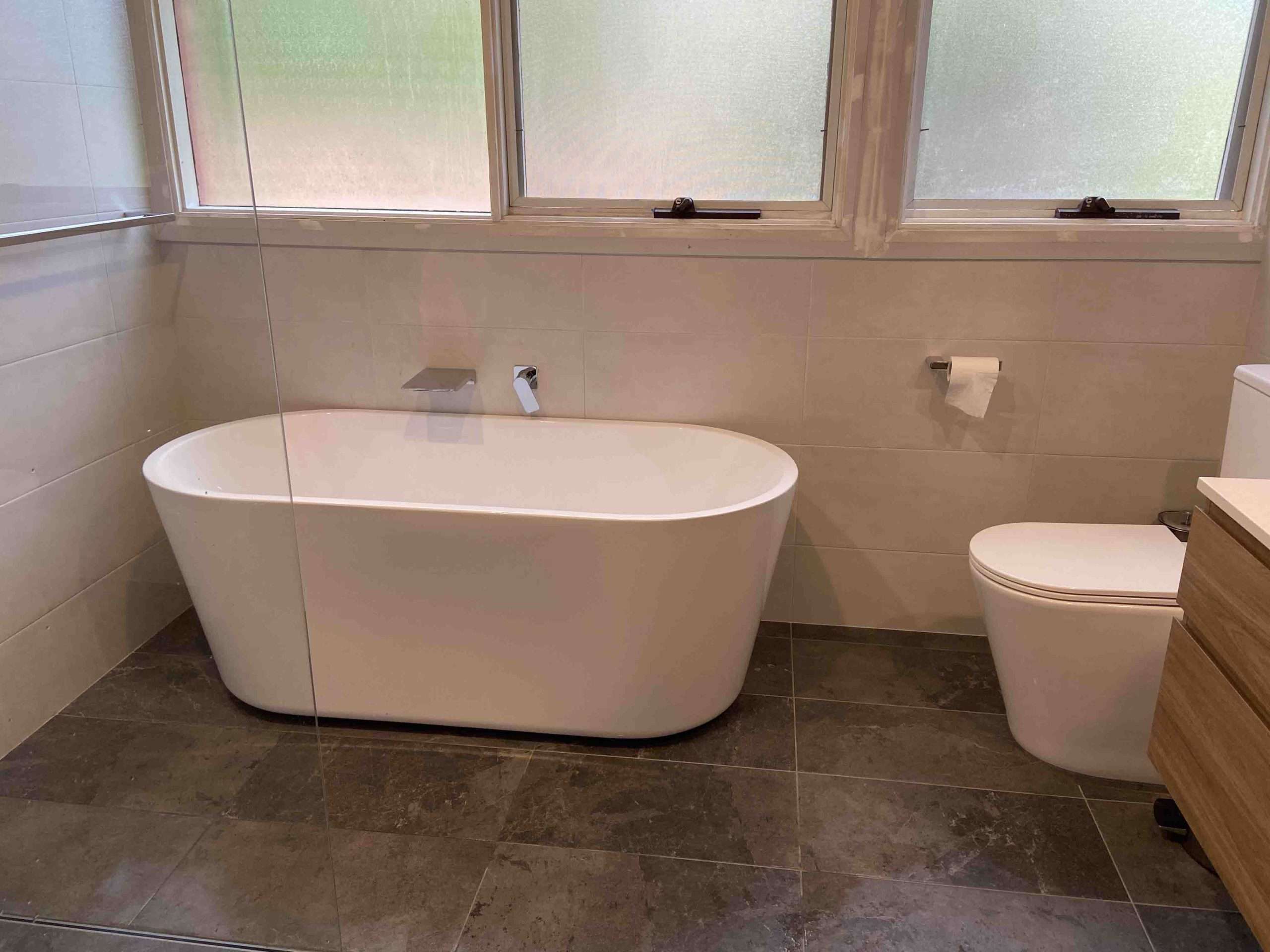 Freestanding bath, toilet and timber vanity on dark floor tiles below frosted windows