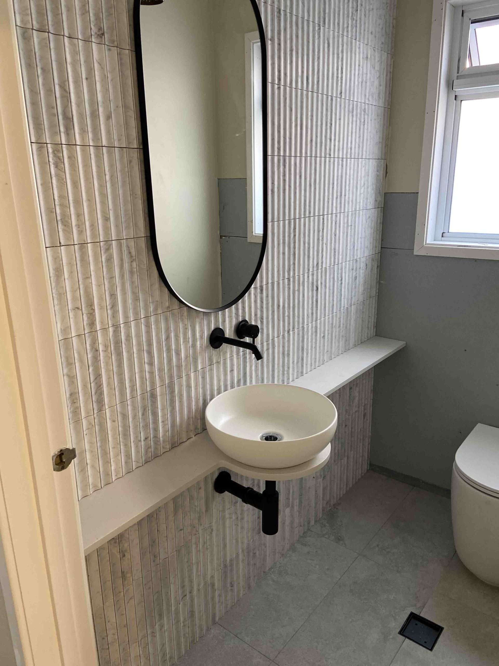 Round basin on a floating shelf below a black-framed oval mirror, with fluted wall tiles