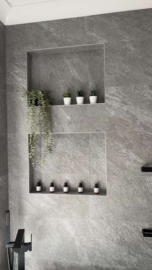 Two recessed niches in a grey-tiled shower holding small plants and bottles, with black tapware