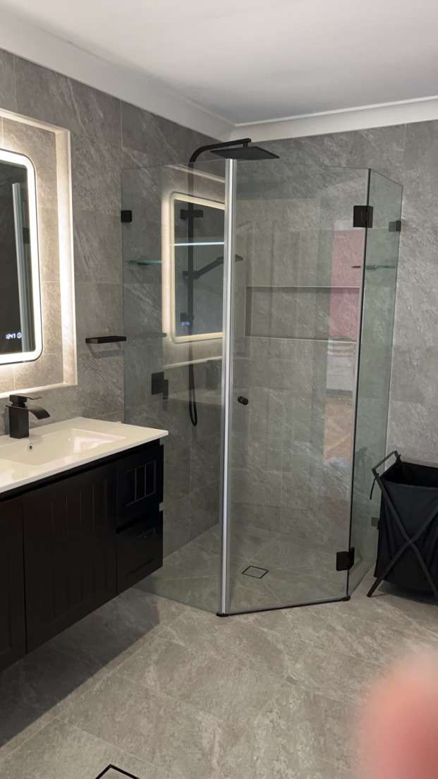 Angled frameless shower screen with black hinges in a grey-tiled bathroom
