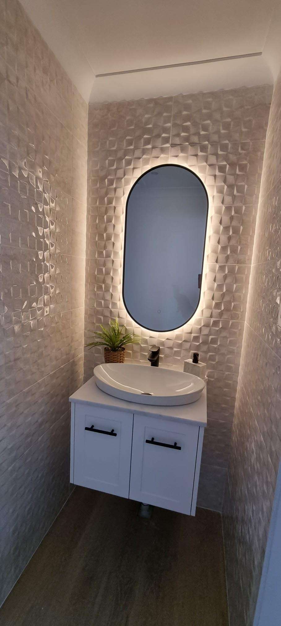 Narrow powder room with a backlit oval mirror, textured tiles and a white wall-hung vanity
