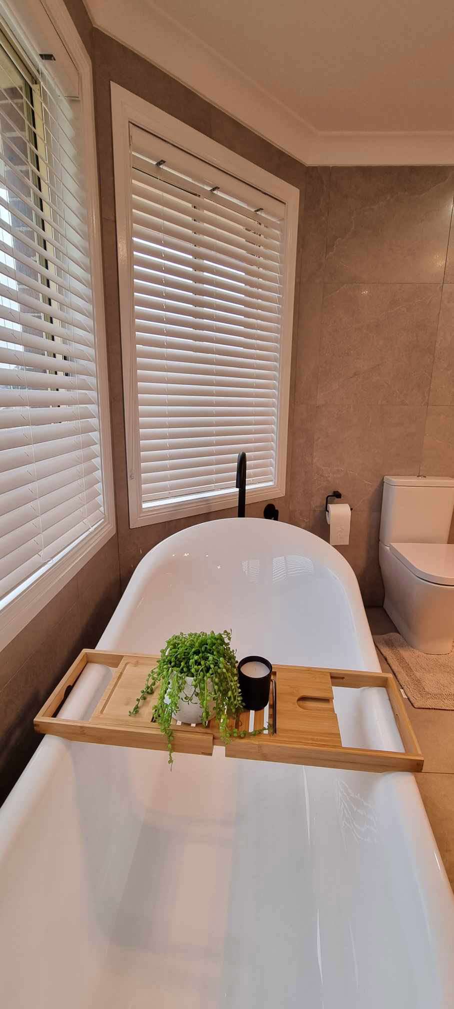Freestanding bath with a bamboo bath caddy holding a plant, beside windows with white blinds