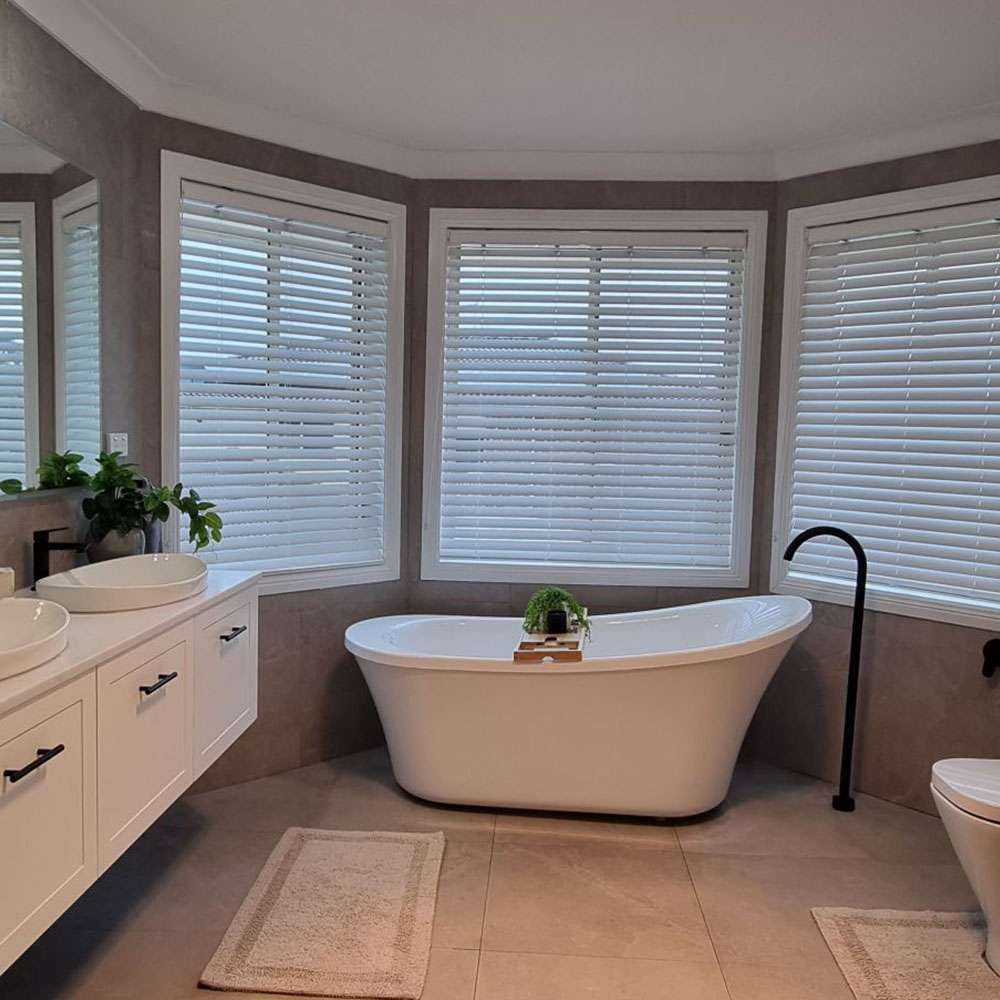 Freestanding bath in a bay window with white blinds, a black floor-mounted tap and white vanity