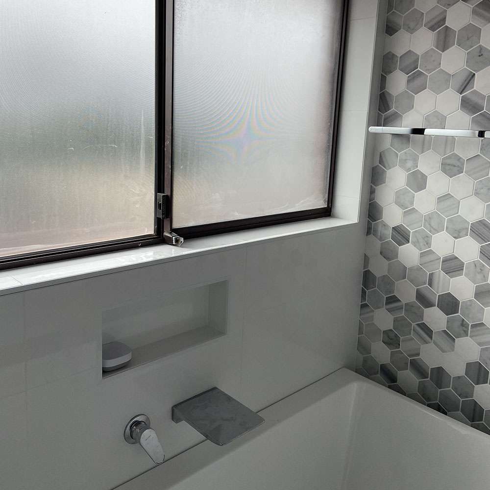 Bath with a grey wall spout below a frosted window, with hexagon mosaic tiles and a niche