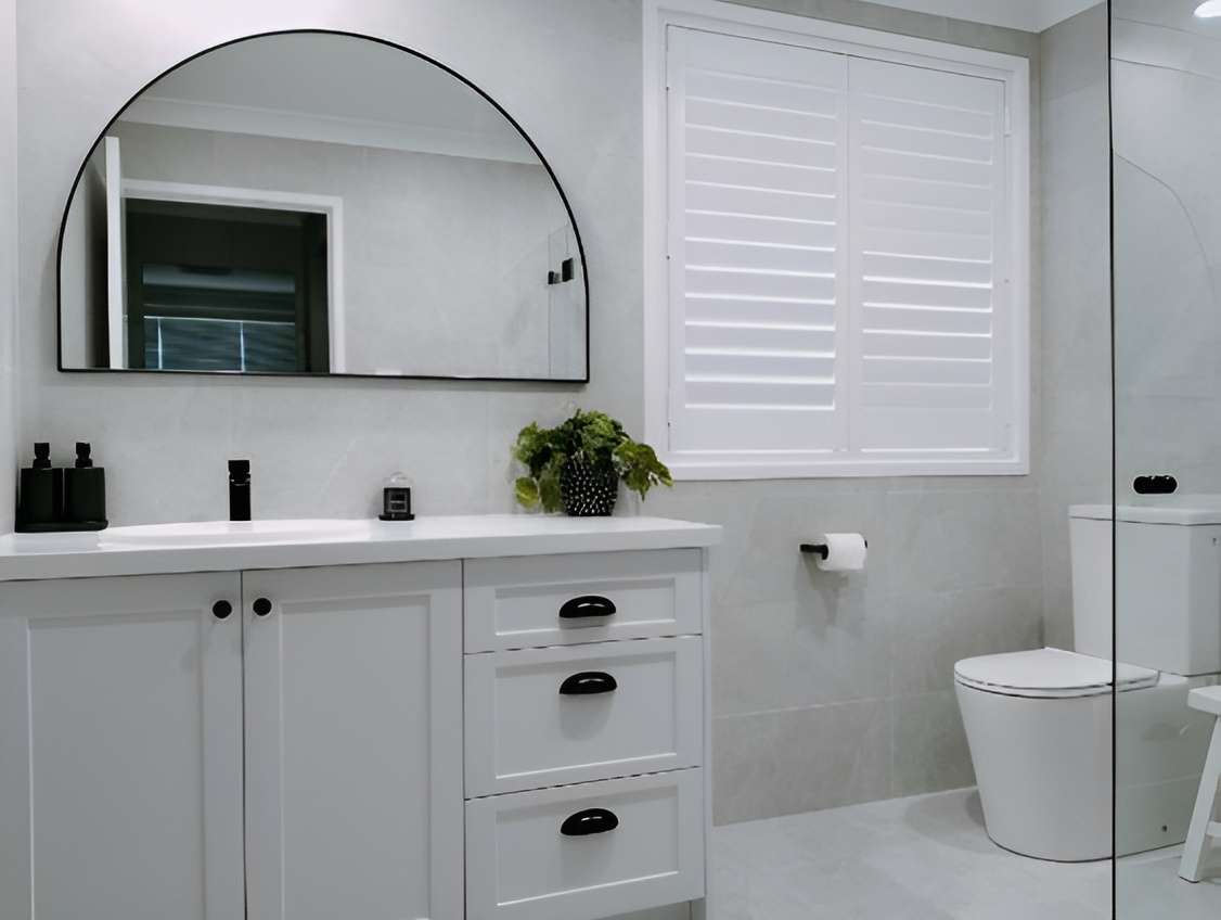 Grey bathroom with a white shaker vanity, black arched mirror, white shutters and toilet