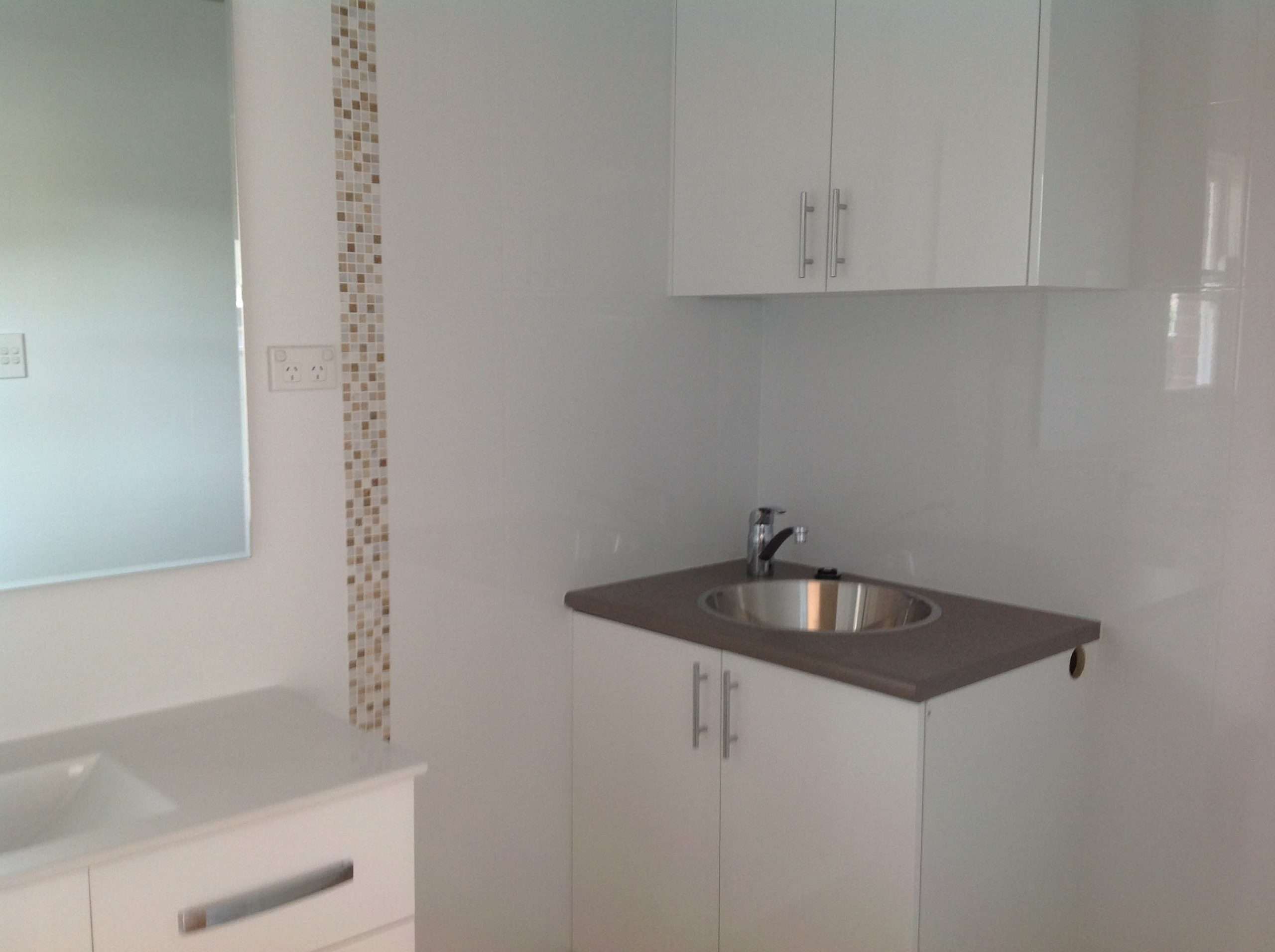 Stainless steel sink in a white cabinet with a grey top, beside a white vanity and mosaic tile strip