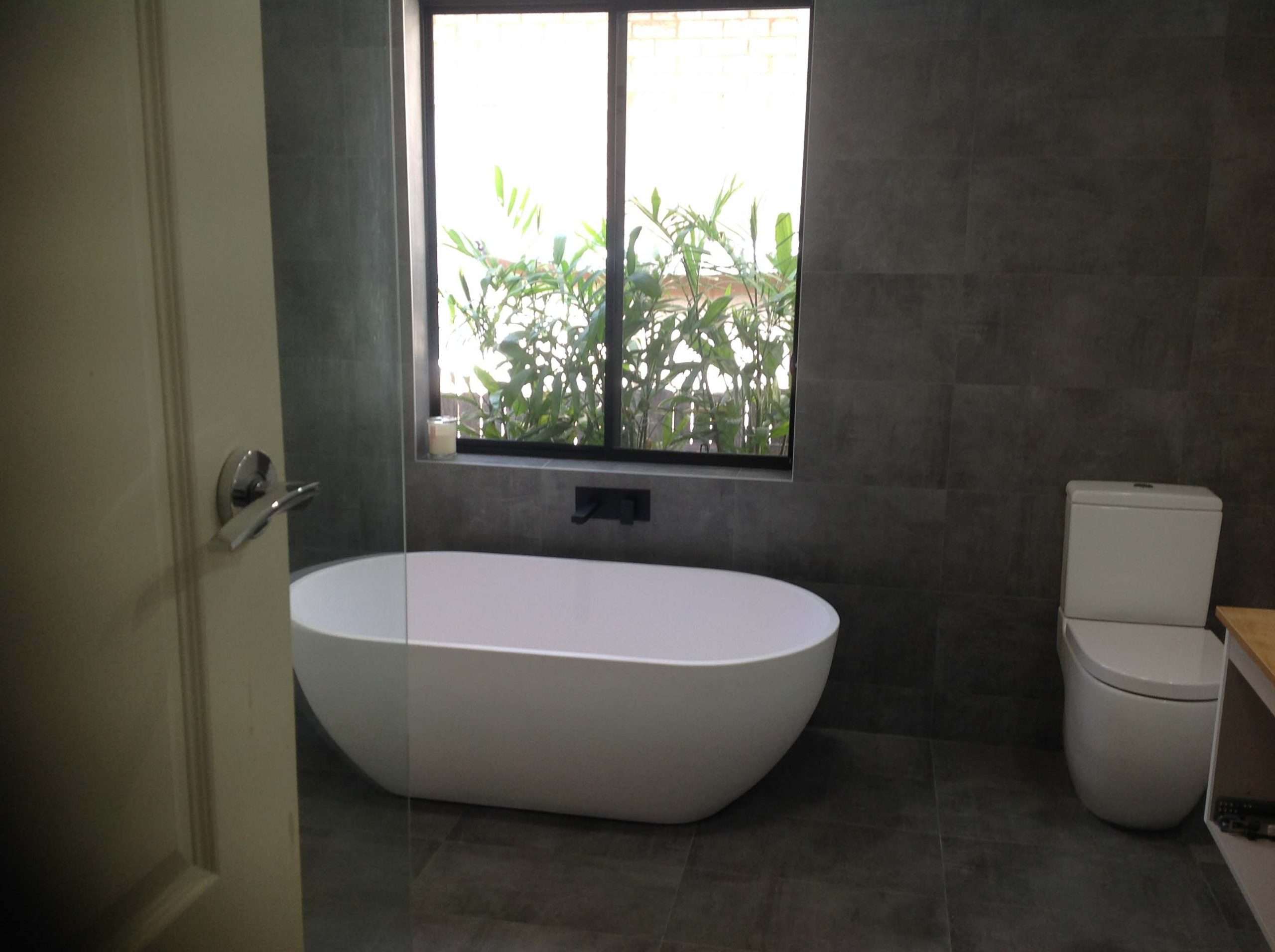 Freestanding bath and toilet in a dark grey tiled bathroom with a large window