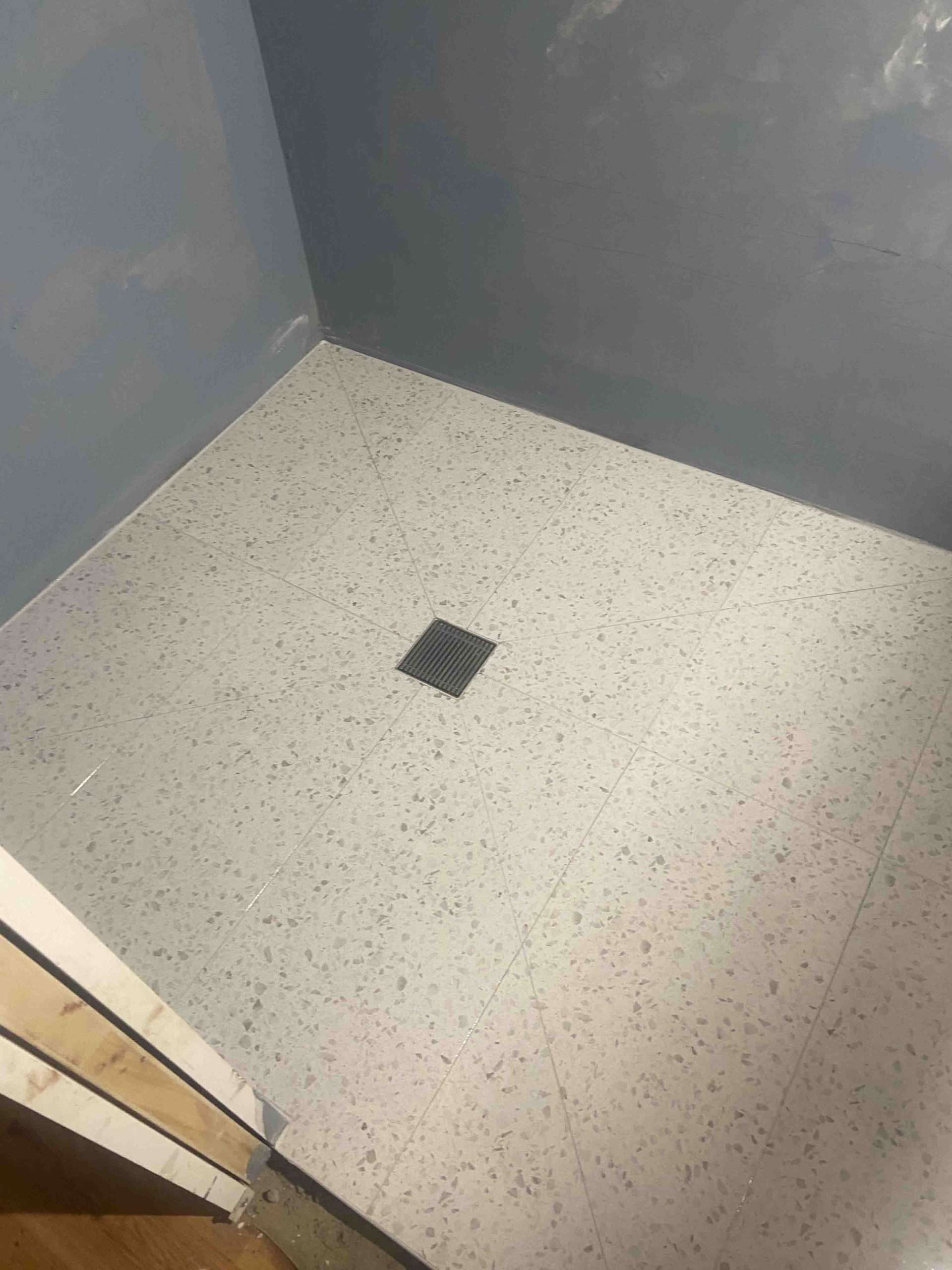 Speckled floor tiles and a square floor waste in a shower with untiled walls