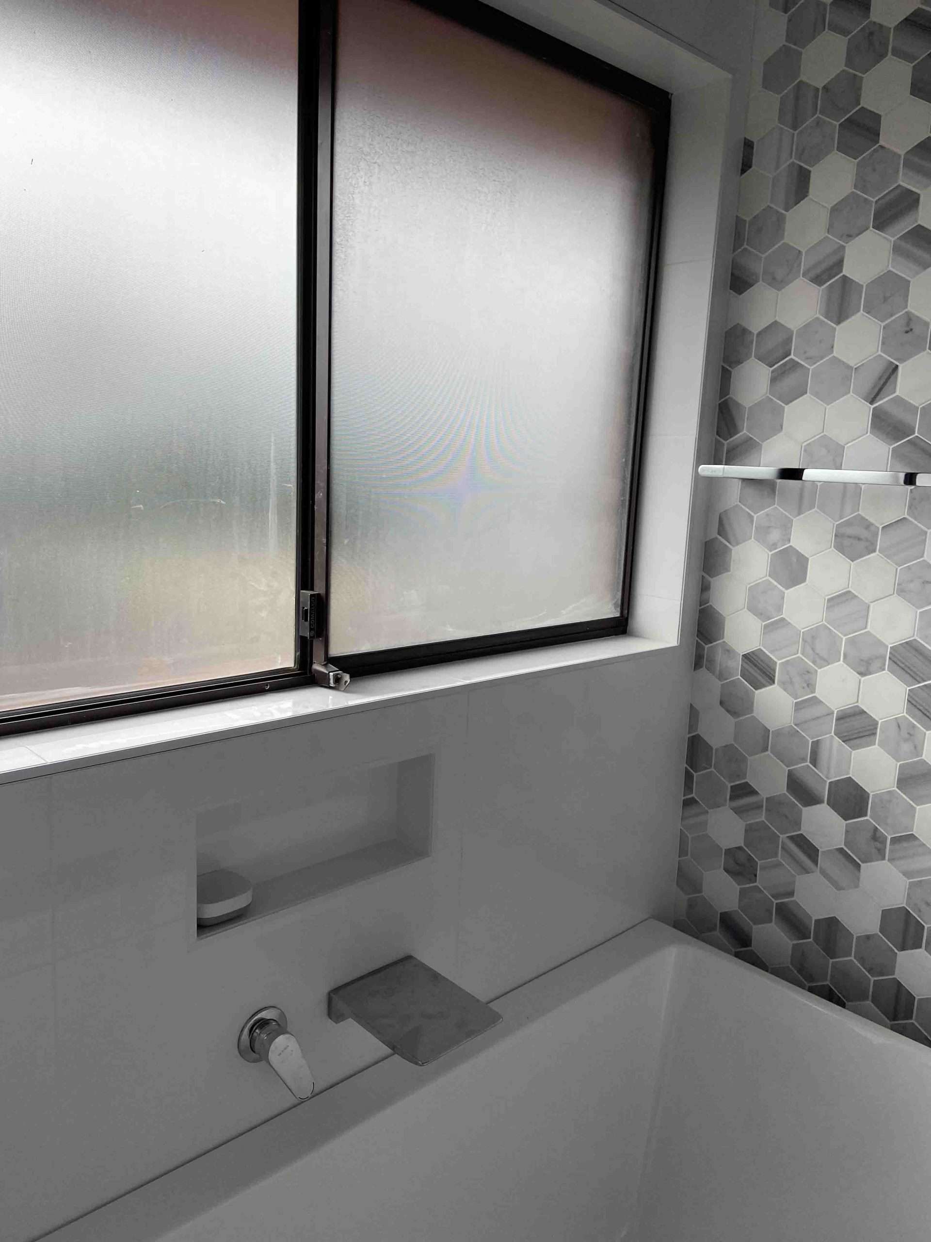 Built-in bath with a wall spout, shower niche and grey hexagon feature tiles below a frosted window